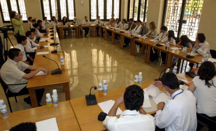 A large group of students sit at multiple long tables facing each other with paper and microphones